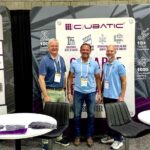 Three professionals posing at the Qubatic booth during a steel detailing industry event, with Qubatic branding and structural steel detailing services displayed in the background.